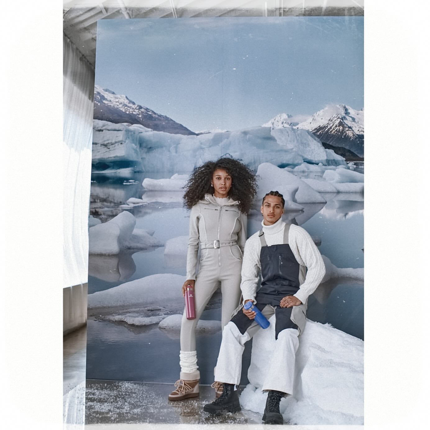 Two models pose in front of a glacier backdrop wearing fashionable winter clothing, including overalls and sweaters. They hold water bottles.