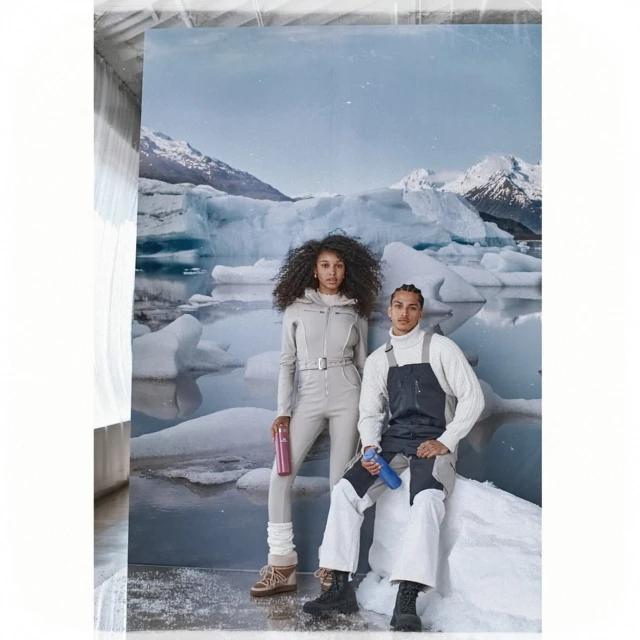Two models pose in front of a glacier backdrop wearing fashionable winter clothing, including overalls and sweaters. They hold water bottles.