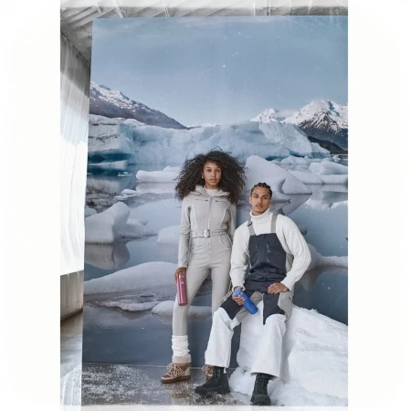 Two models pose in front of a glacier backdrop wearing fashionable winter clothing, including overalls and sweaters. They hold water bottles.