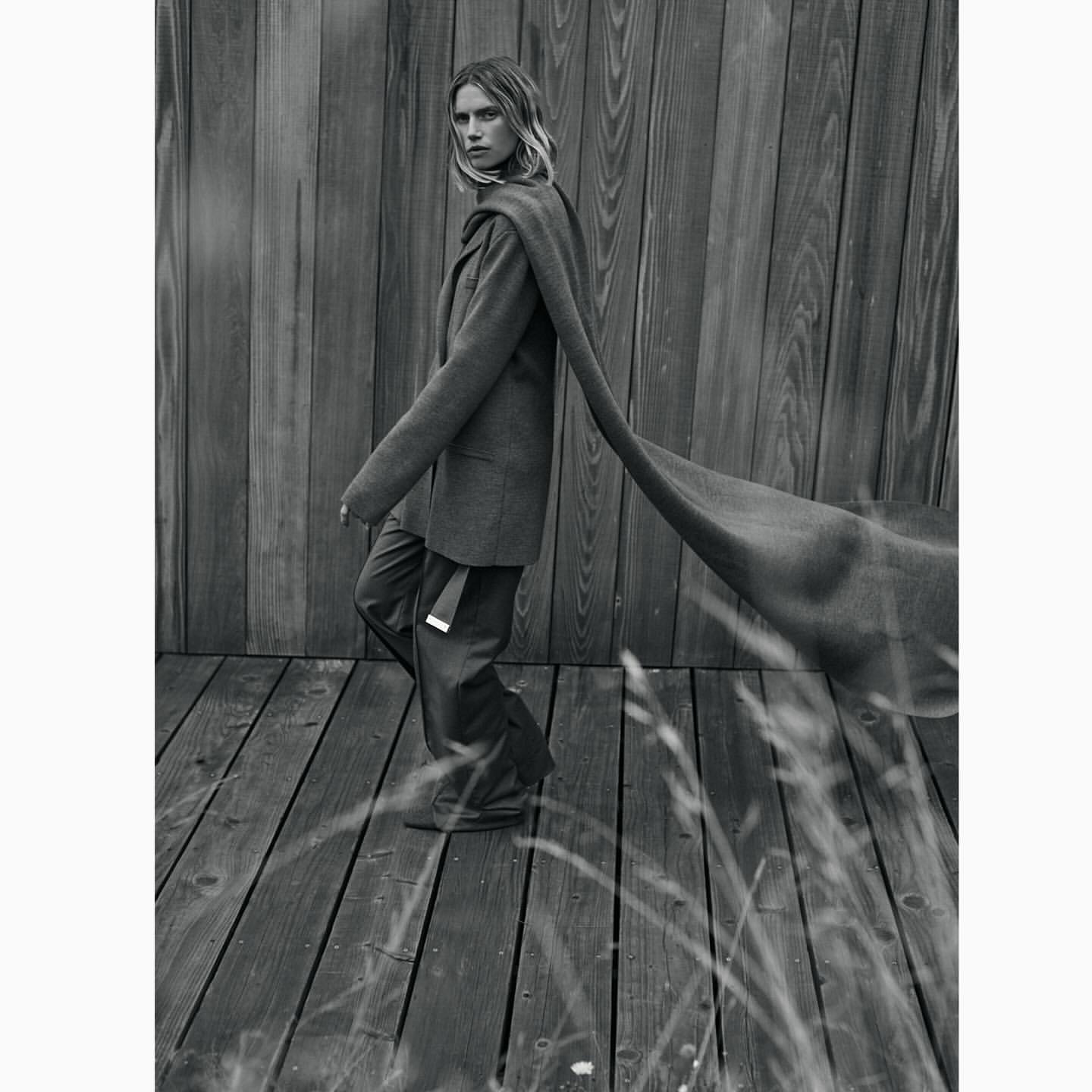 Monochrome portrait of a woman wearing a tailored blazer and long scarf standing on a wooden deck by a wooden wall.