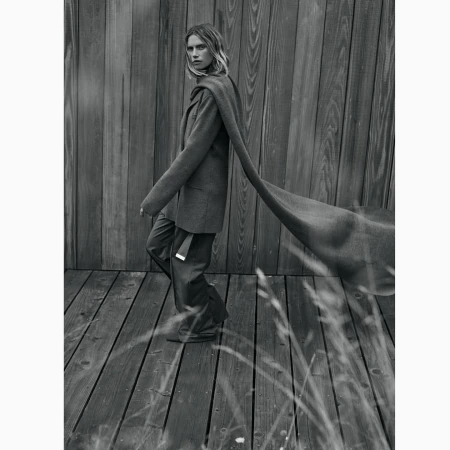 Monochrome portrait of a woman wearing a tailored blazer and long scarf standing on a wooden deck by a wooden wall.