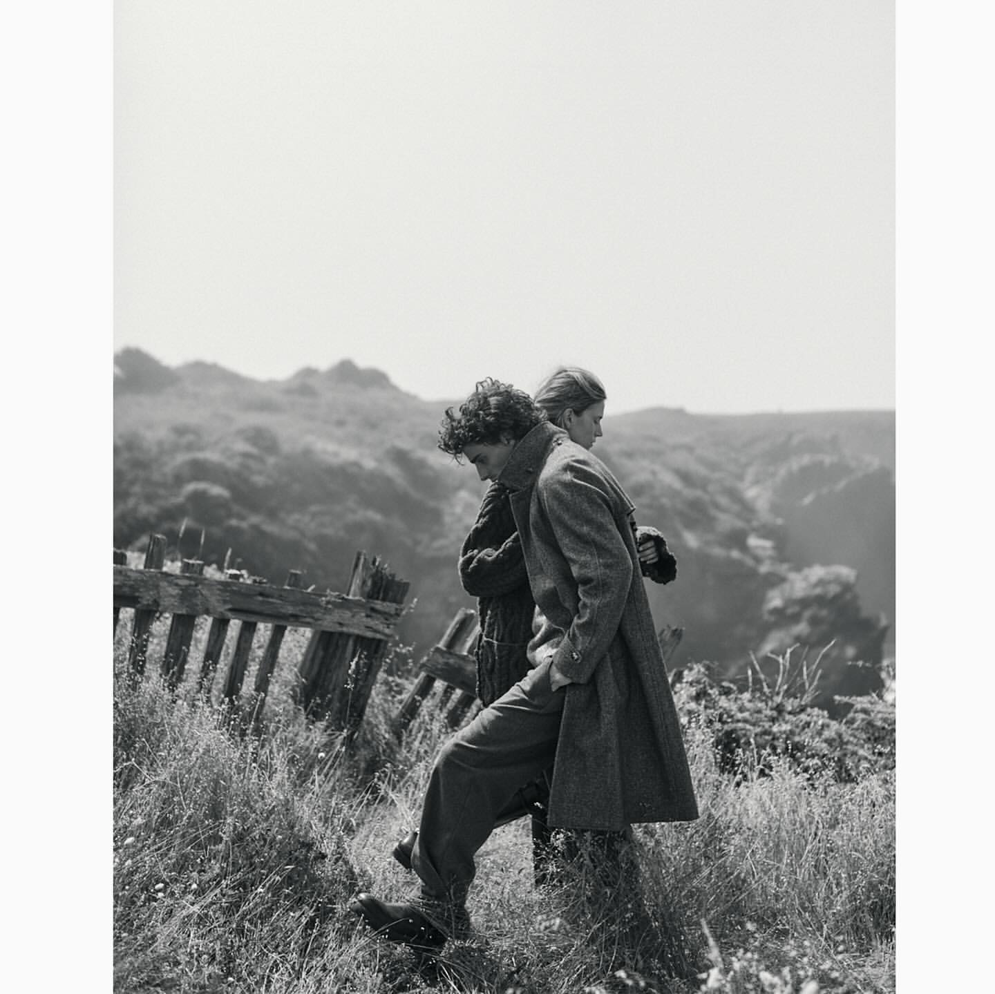 Two people in coats walking along a grassy hill next to a weathered fence, with a blurred landscape in the background, black and white.