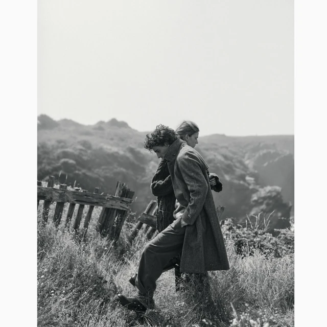 Two people in coats walking along a grassy hill next to a weathered fence, with a blurred landscape in the background, black and white.