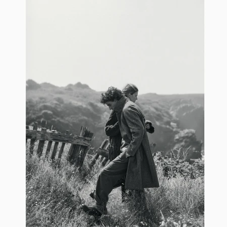 Two people in coats walking along a grassy hill next to a weathered fence, with a blurred landscape in the background, black and white.