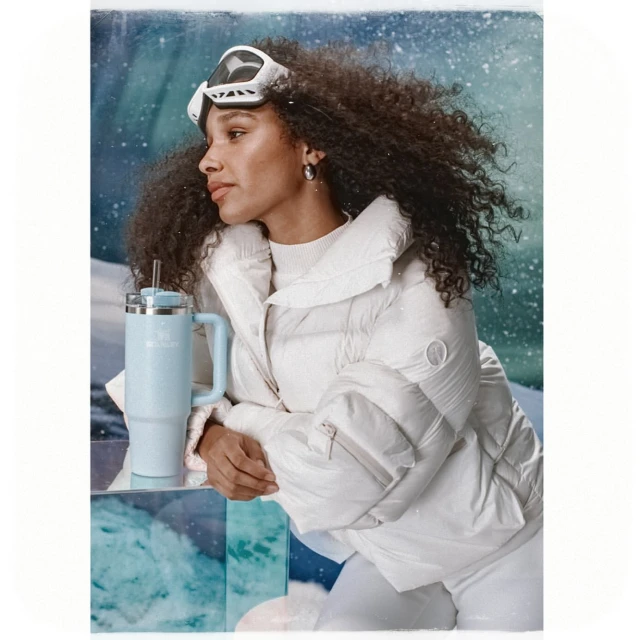 A woman in a white puffer jacket with ski goggles poses with a light blue Stanley cup in a winter scene with snowy backgrounds.