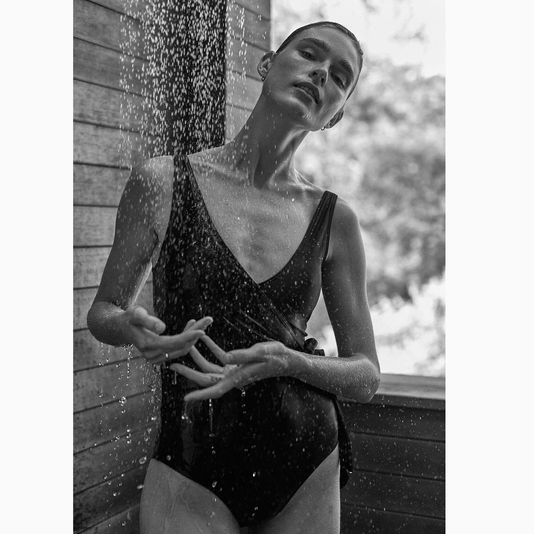 Black-and-white portrait of a woman in a dark swimsuit standing under an outdoor shower with wet hair and water droplets.