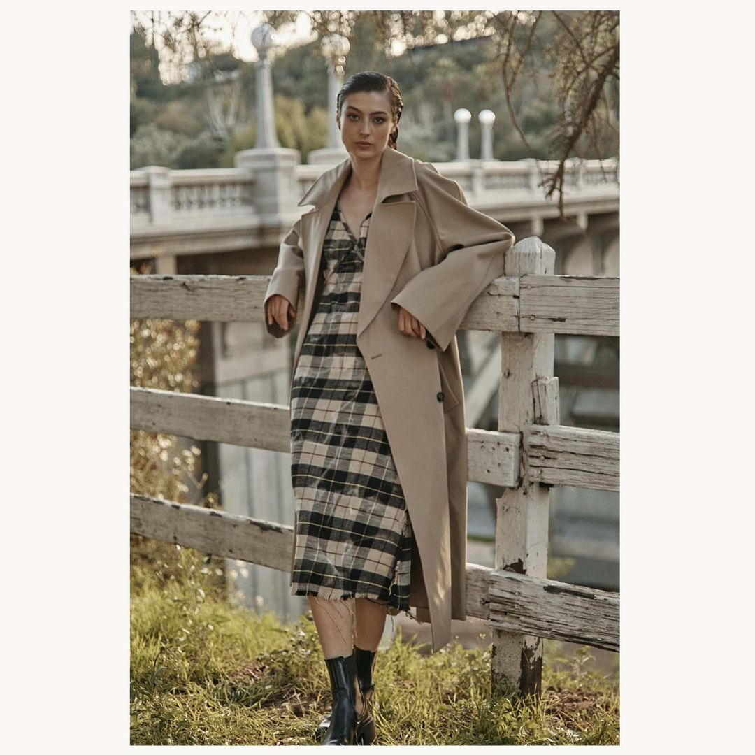 A fashionable woman wearing a trench coat and plaid dress leans against a white fence, set against an outdoor backdrop.