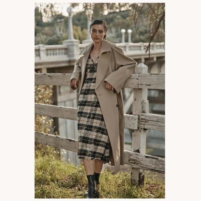 A fashionable woman wearing a trench coat and plaid dress leans against a white fence, set against an outdoor backdrop.