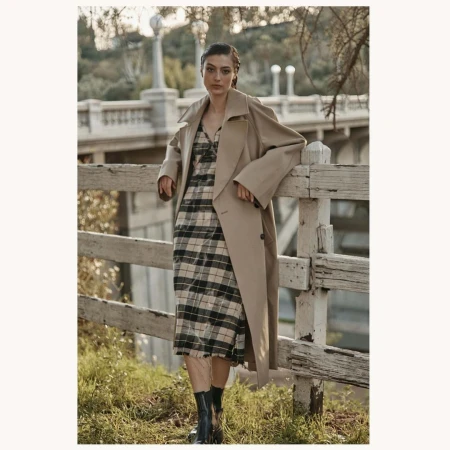 A fashionable woman wearing a trench coat and plaid dress leans against a white fence, set against an outdoor backdrop.