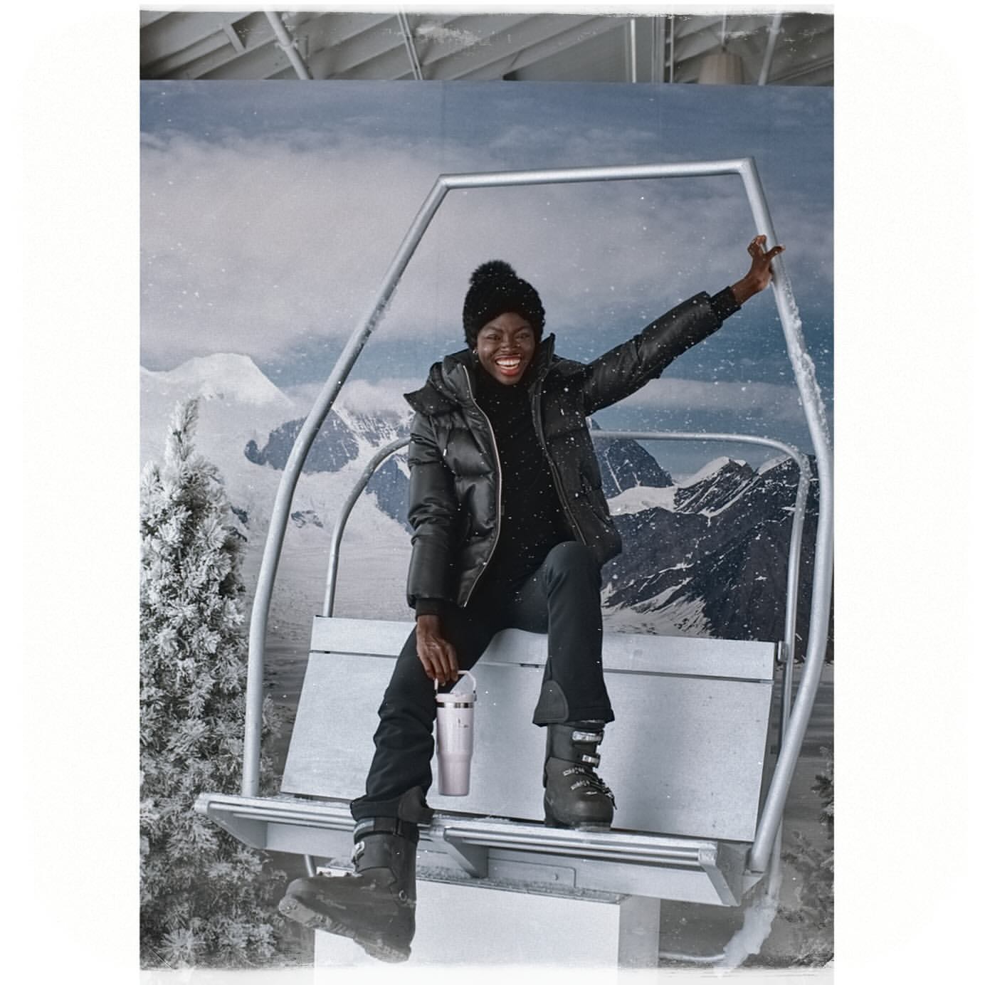A happy Black woman in a black puffer jacket, black ski pants, and boots smiles while seated on a ski lift against a snowy mountain backdrop.