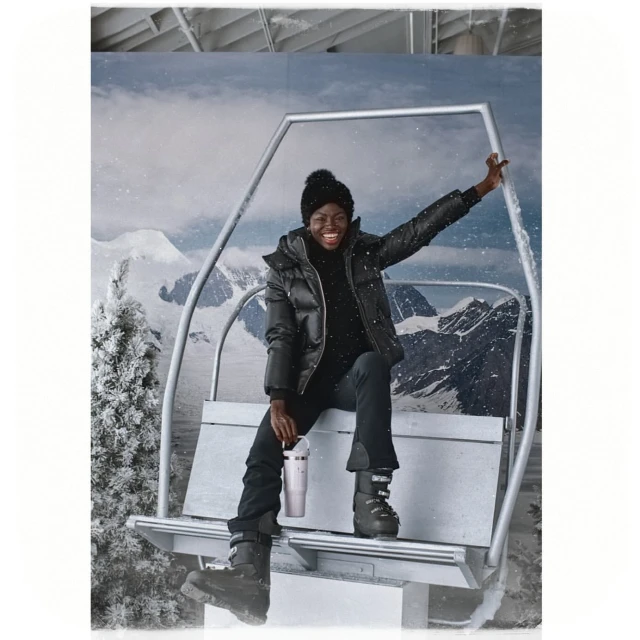 A happy Black woman in a black puffer jacket, black ski pants, and boots smiles while seated on a ski lift against a snowy mountain backdrop.