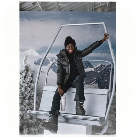 A happy Black woman in a black puffer jacket, black ski pants, and boots smiles while seated on a ski lift against a snowy mountain backdrop.