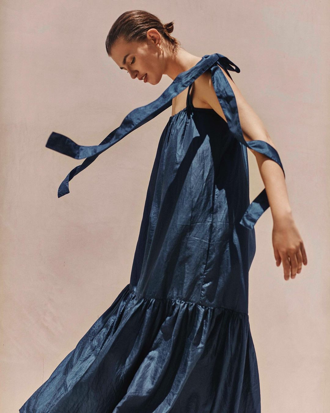 Elegant woman in a flowing navy blue dress with shoulder ties, captured in a soft studio light against a neutral backdrop for a fashion editorial.