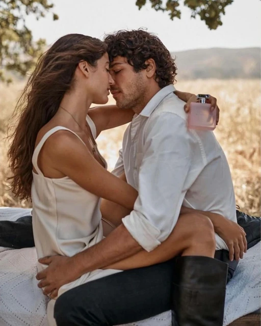 A romantic couple embrace outdoors in warm light, with a perfume bottle visible in the background, conveying an intimate, sunlit moment.