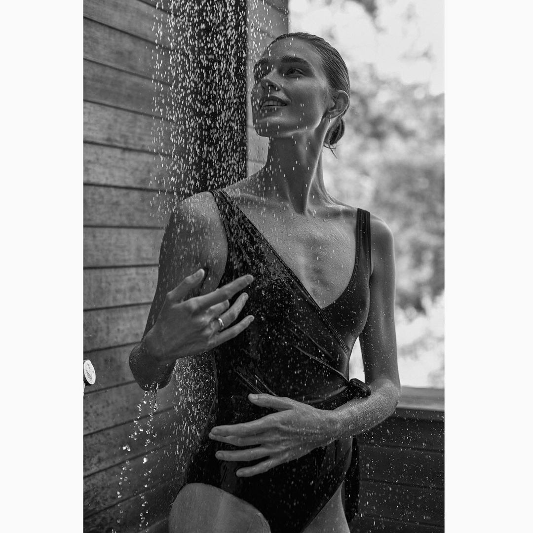 Black and white portrait of a woman in a dark swimsuit standing under a shower with a wood paneled wall and greenery visible.