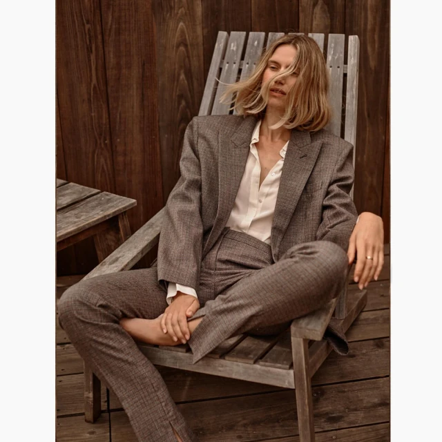 Blonde woman poses in a brown suit on a wooden chair outdoors. She wears a white shirt, a brown plaid blazer and trousers, and sits casually.