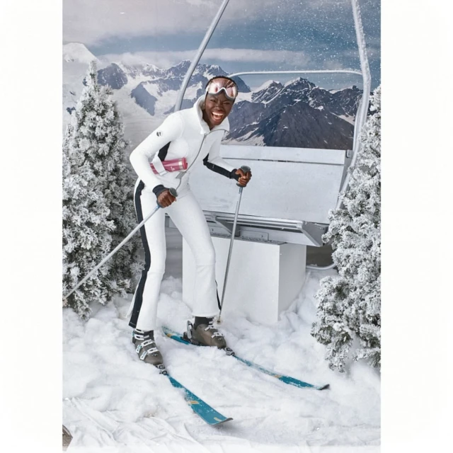 Adult woman poses on skis with ski poles in snow wearing a white ski suit, goggles, and boots, with snow-covered trees and mountains.