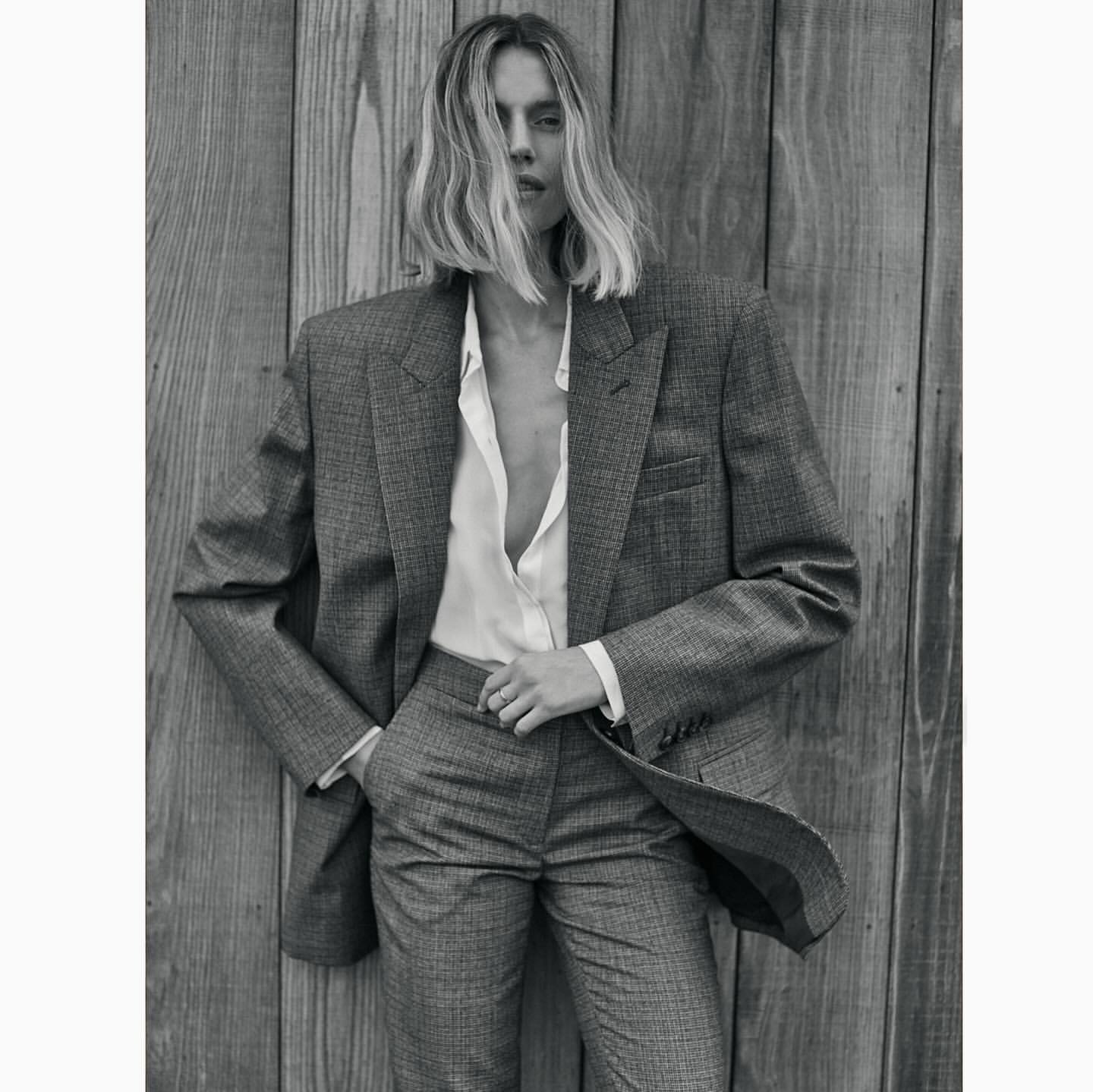 Black and white portrait of an adult female in a grey pantsuit leaning against a wooden wall, capturing a classic fashion editorial.
