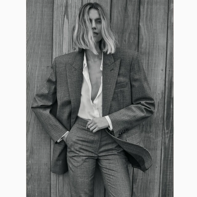 Black and white portrait of an adult female in a grey pantsuit leaning against a wooden wall, capturing a classic fashion editorial.