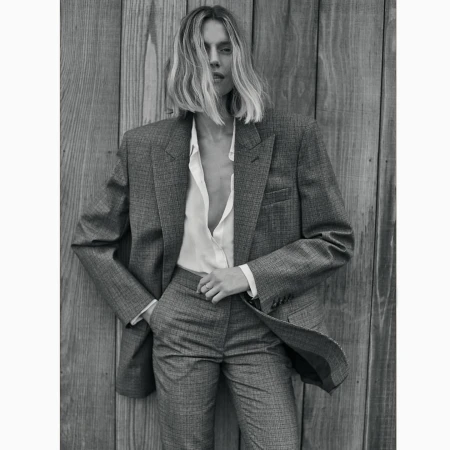 Black and white portrait of an adult female in a grey pantsuit leaning against a wooden wall, capturing a classic fashion editorial.