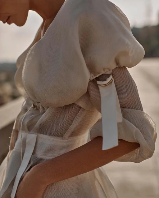 Close-up of a fashionably dressed person in a puffy-sleeved, light-colored dress outdoors with a neutral background. Beauty shot, eye-level.