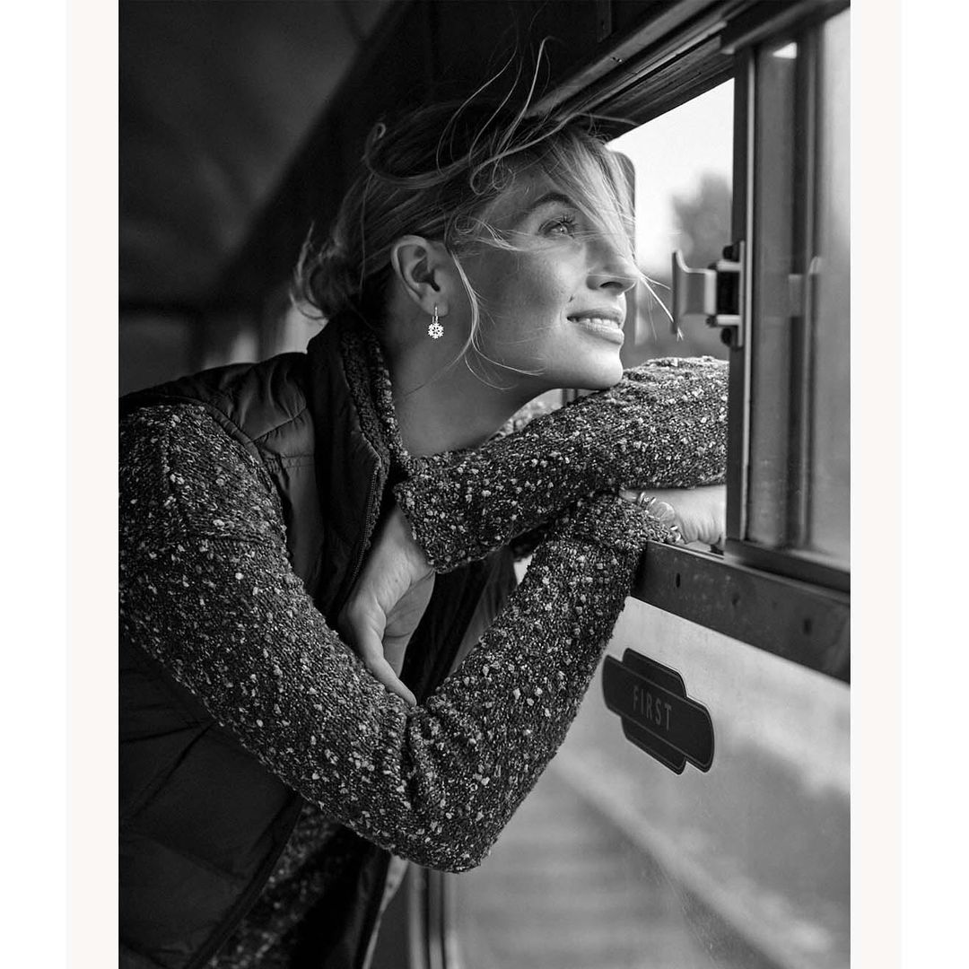 Black and white portrait of a fair-skinned woman gazing out a train window with a slight smile, wearing a vest and patterned sweater.