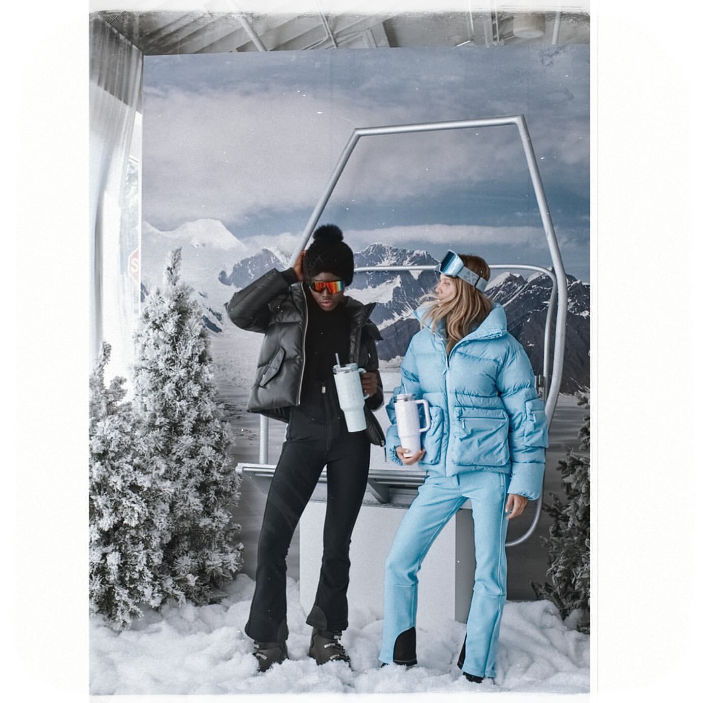 Two women in winter fashion stand in a staged ski lift scene with snow-covered trees and mountain backdrop, holding travel mugs.
