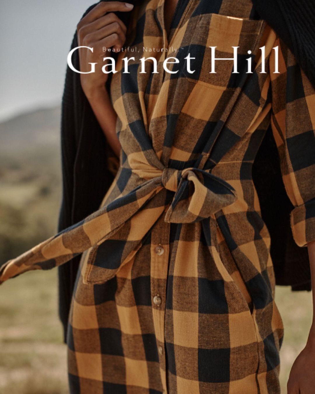 A woman in a black and tan plaid dress with a tie around the waist stands outdoors, showcasing fashion and style in a medium shot.