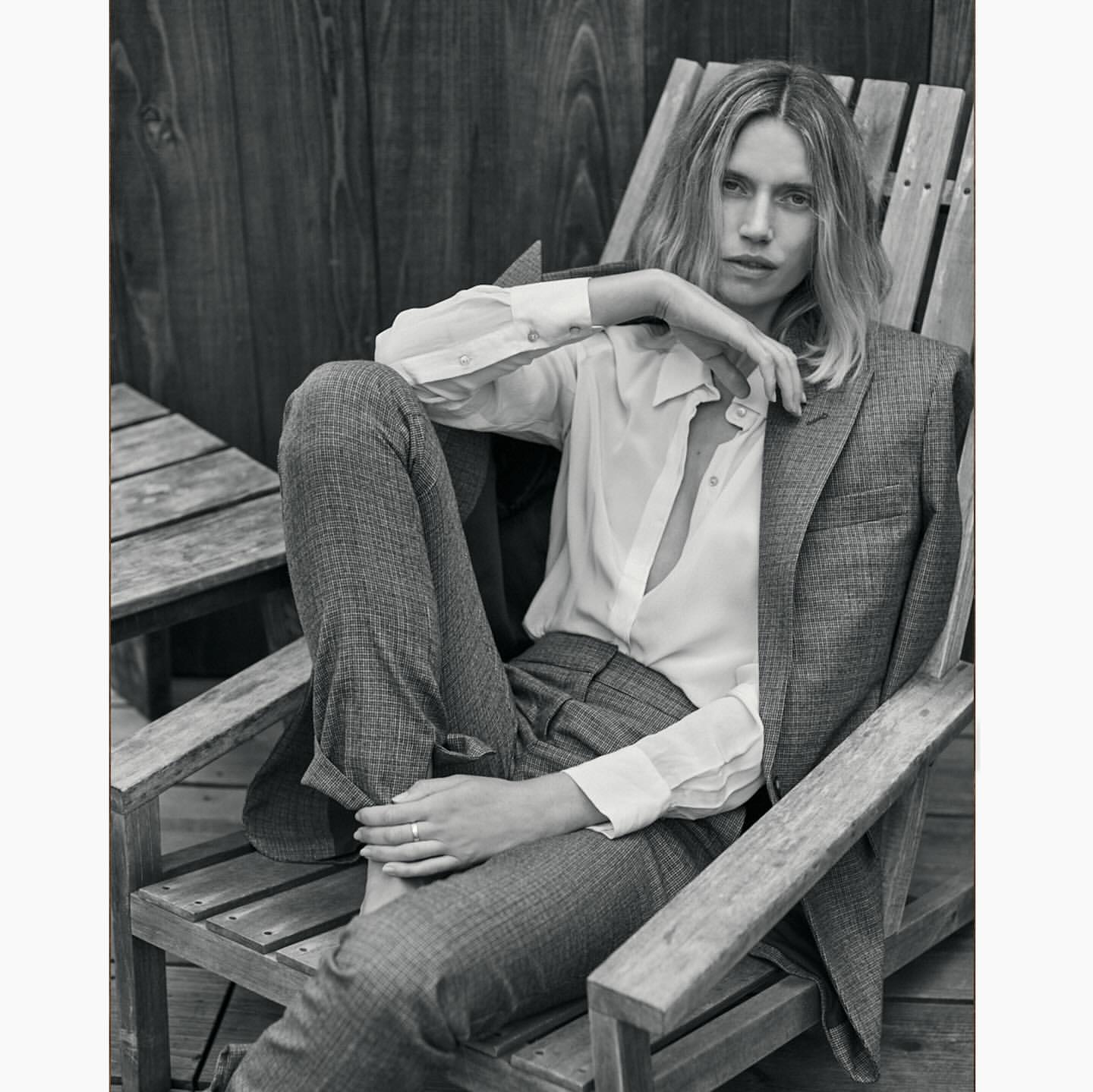 Monochrome portrait of a blonde woman in a blazer and white button-down shirt, sitting on a wooden chair outdoors.