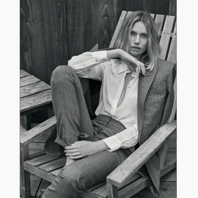 Monochrome portrait of a blonde woman in a blazer and white button-down shirt, sitting on a wooden chair outdoors.