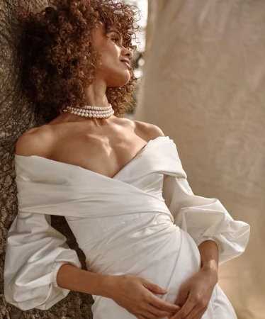 Woman with curly hair wearing a white off-shoulder dress and pearl choker leaning against a tree trunk in Los Angeles.