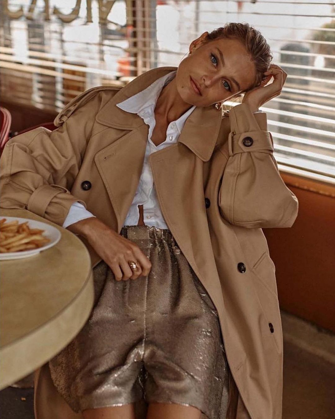 Blonde woman poses indoors in a tan trench coat and gold shorts at a table with fries, illuminated by natural window light.