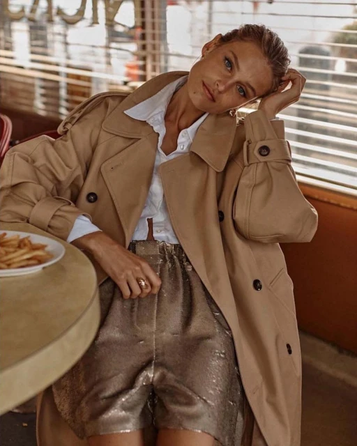 Blonde woman poses indoors in a tan trench coat and gold shorts at a table with fries, illuminated by natural window light.