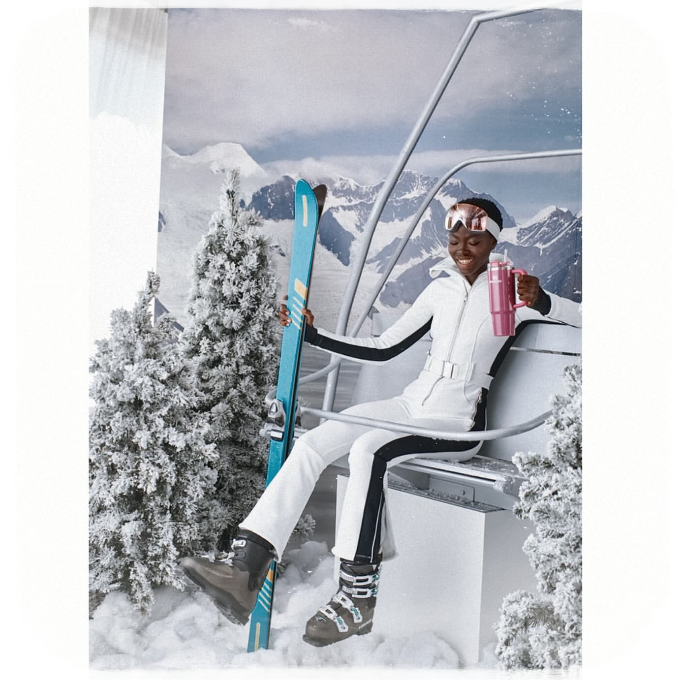 Woman in ski outfit on a ski lift with skis, wearing goggles and holding a thermos against a snowy mountain backdrop.