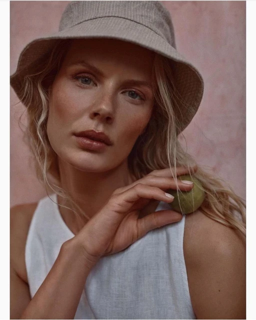 Close-up of a blonde woman with a bucket hat, wearing a white top, holding a green stone, with a textured pink backdrop. Beauty shot.