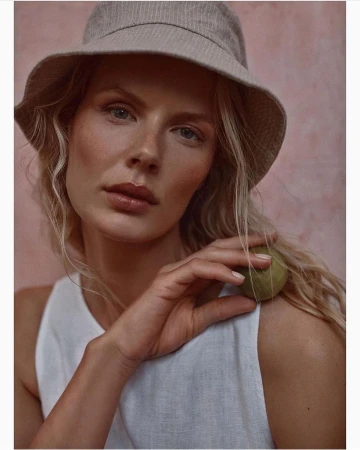 Close-up of a blonde woman with a bucket hat, wearing a white top, holding a green stone, with a textured pink backdrop. Beauty shot.