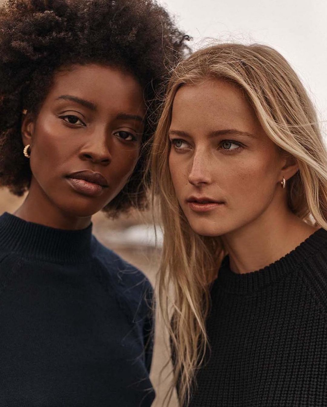 Portrait of two diverse women with an outdoor setting, wearing dark sweaters and natural hair. Fashion or beauty editorial style image.
