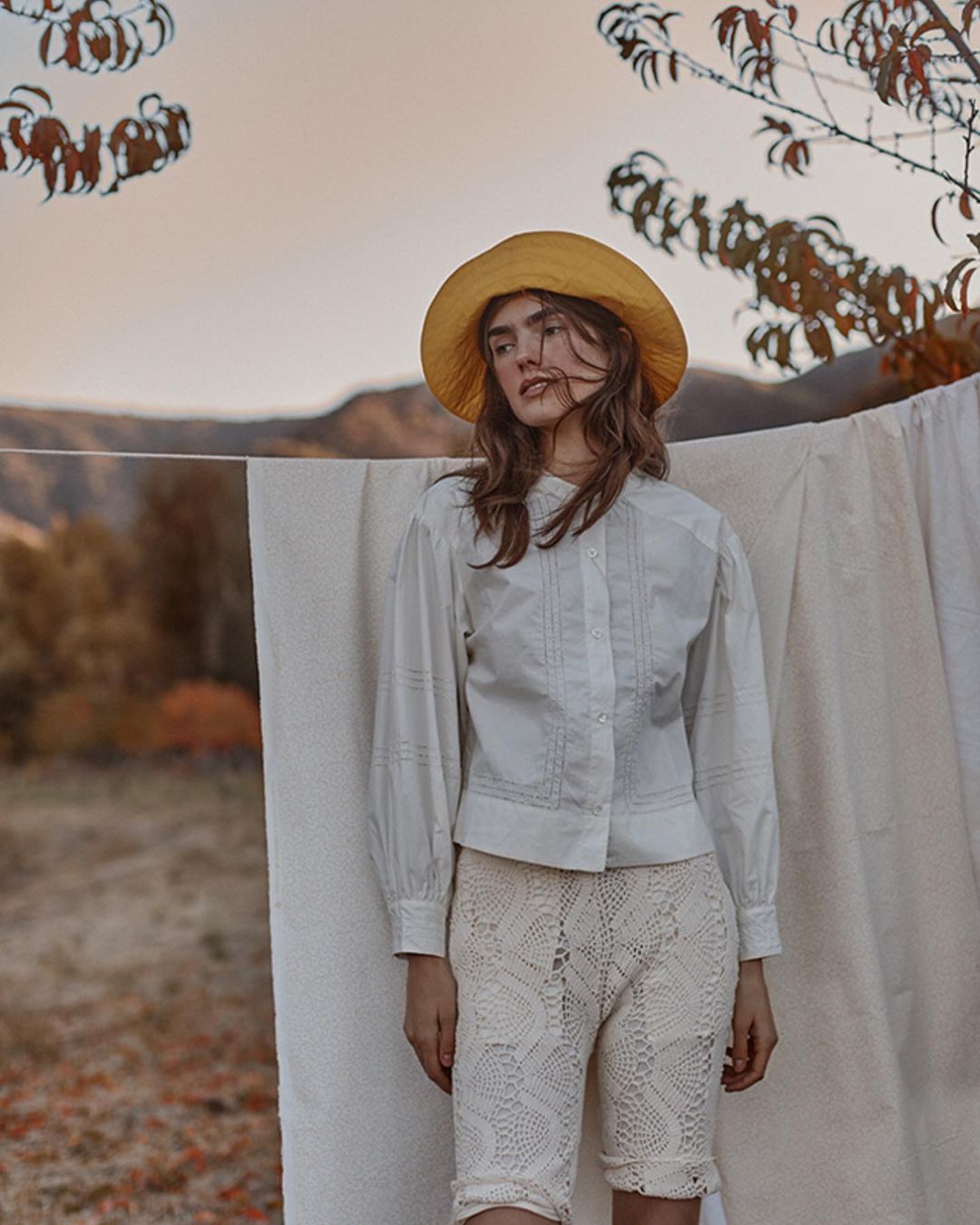 Young woman poses in white blouse and crocheted shorts with straw hat in a rural setting, showcasing fashion and natural light.