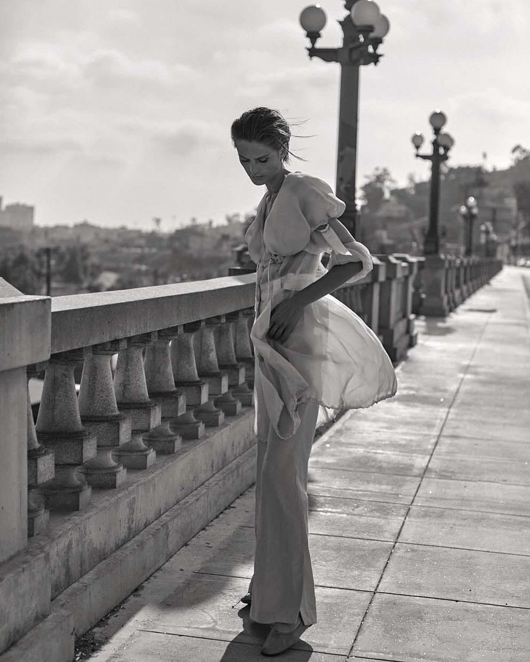 Black and white fashion shot of an adult woman wearing a long dress, standing on a bridge with an urban background and lamp posts.