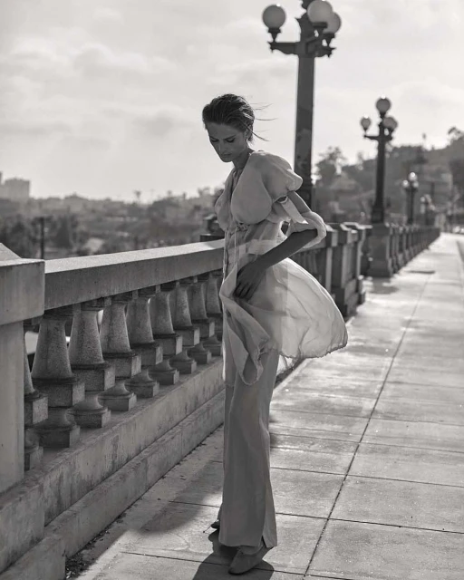 Black and white fashion shot of an adult woman wearing a long dress, standing on a bridge with an urban background and lamp posts.