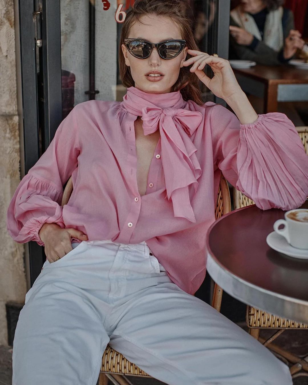 Elegant woman in pink blouse and white pants sits at cafe table with coffee, posing in diffused daylight with neutral expression.