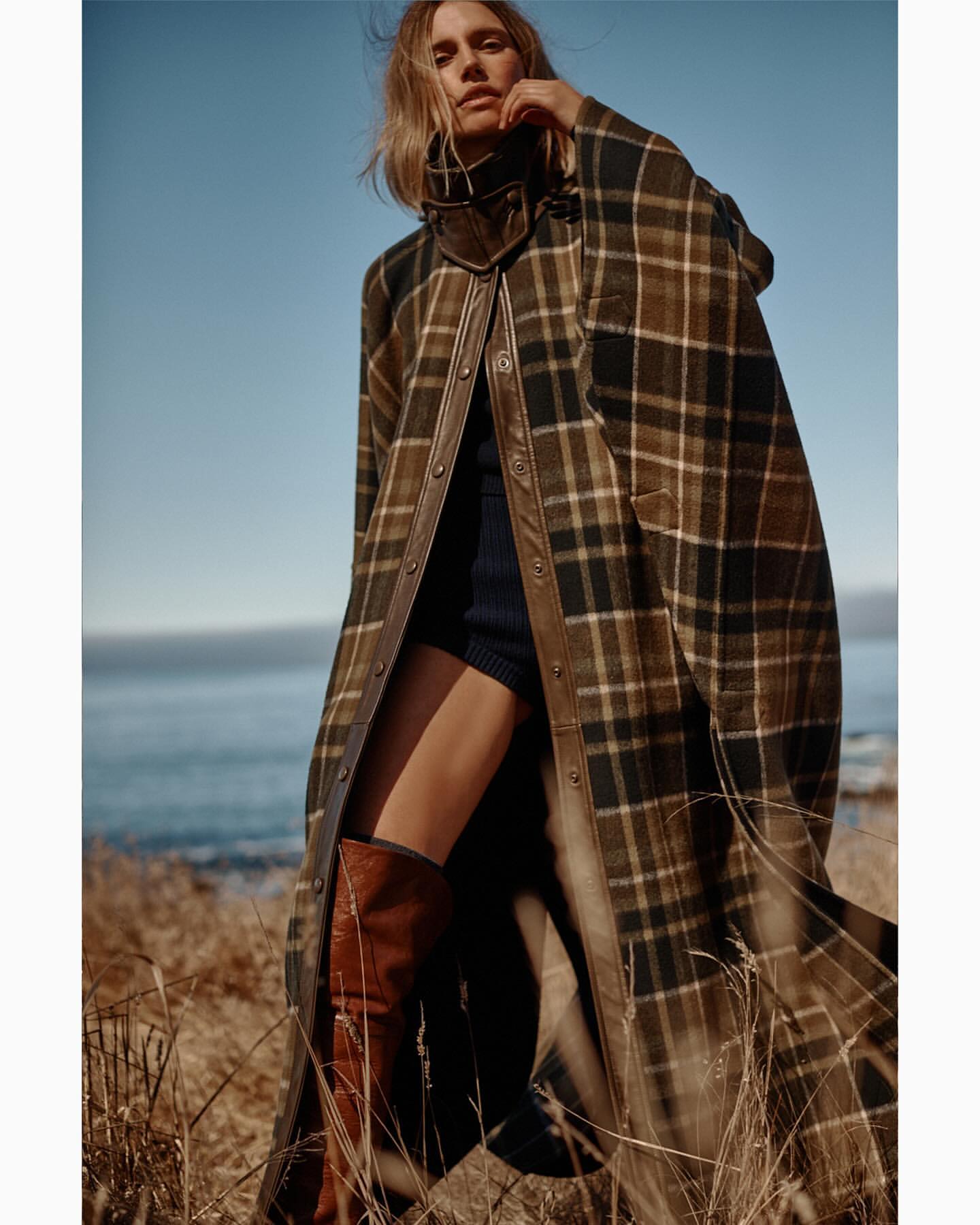 Woman wearing a plaid poncho with leather trim and brown boots stands in a field of dry grass with a view of the ocean and sky.