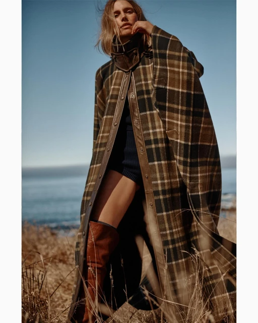 Woman wearing a plaid poncho with leather trim and brown boots stands in a field of dry grass with a view of the ocean and sky.