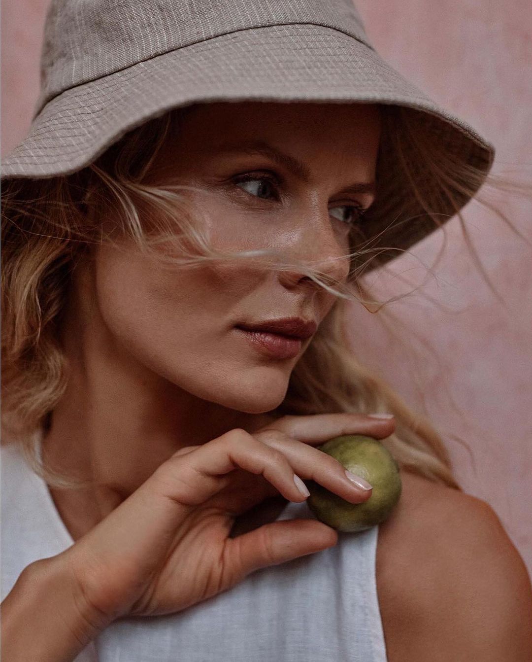 Woman wearing a bucket hat holds a green sphere against a pink background in a close-up shot emphasizing textures and neutral tones.