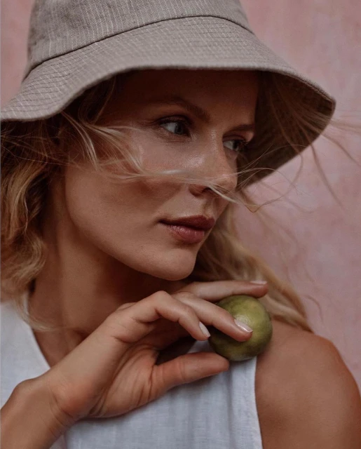 Woman wearing a bucket hat holds a green sphere against a pink background in a close-up shot emphasizing textures and neutral tones.