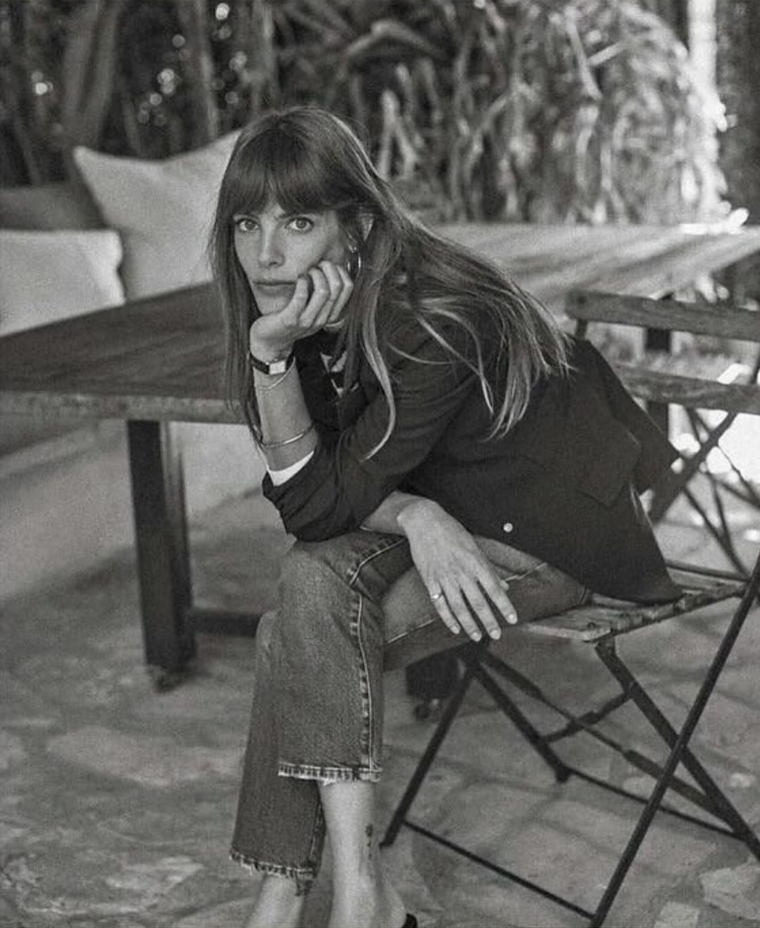 Black and white portrait of a woman sitting on a wooden chair outdoors with her chin resting on her hand.