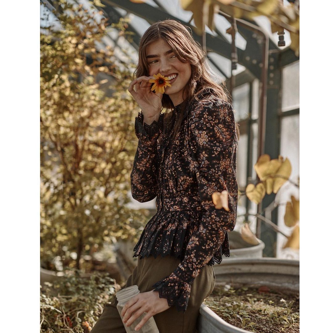 Smiling woman in floral print blouse with lace trim poses with a yellow flower inside a greenhouse with plants and natural light.