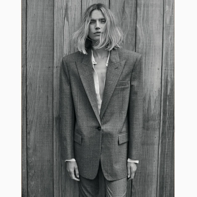 Woman in an oversized gray suit stands in front of a wooden plank wall in a studio, showcasing a blend of fashion and portraiture.