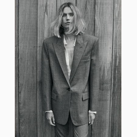 Woman in an oversized gray suit stands in front of a wooden plank wall in a studio, showcasing a blend of fashion and portraiture.