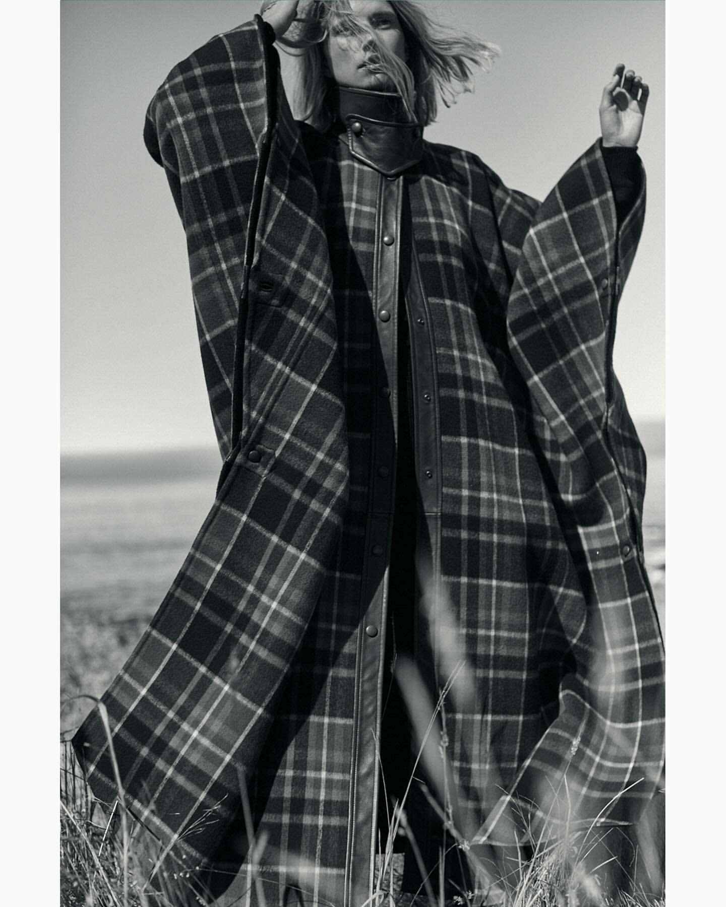 Monochrome portrait of a fair-skinned model in a plaid poncho-style coat. Tall grass is in the foreground, ocean and sky in the background.